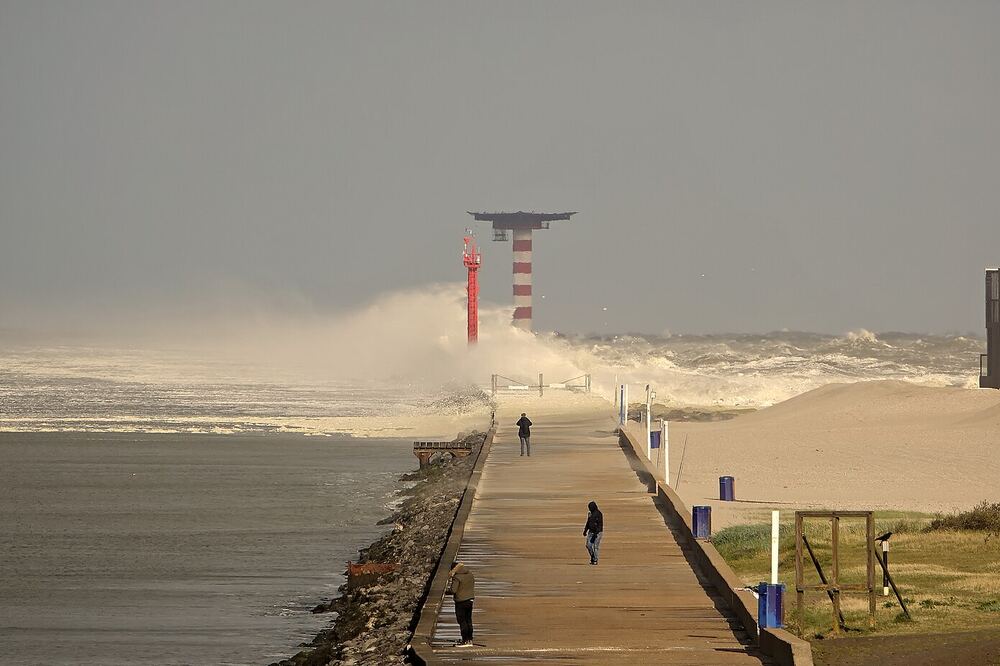 Wandelaars op de pier bij Hoek van Holland terwijl een golf uiteenslaat naast de rood-witte lichttoren, met strand en duinen aan de rechterkant