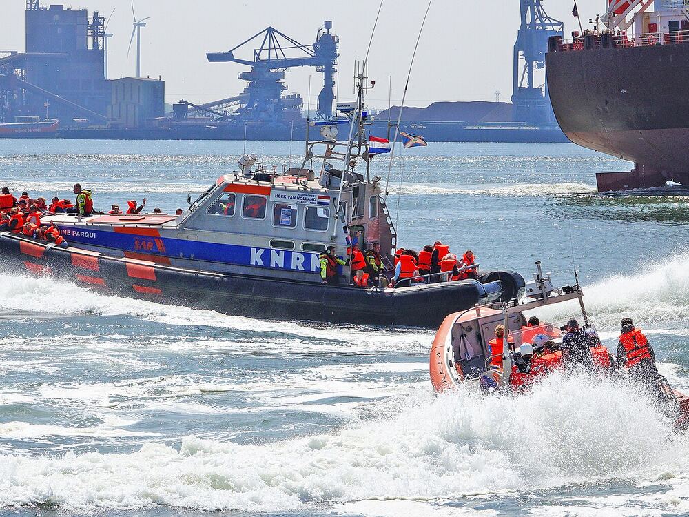 Reddingboot vol opvarenden in oranje reddingvesten vaart tijdens een open dag door de havenmond bij Hoek van Holland, met havenkranen op de achtergrond